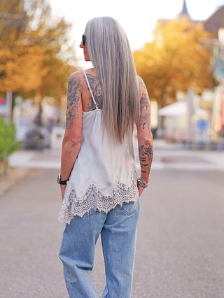 Person von hinten in einer ruhigen Straße, trägt das OFF#DLY Top Violetta champagner mit Spitzenbesatz am Saum, kombiniert mit hellen Jeans. Tattoos und Armbänder ergänzen den Look.