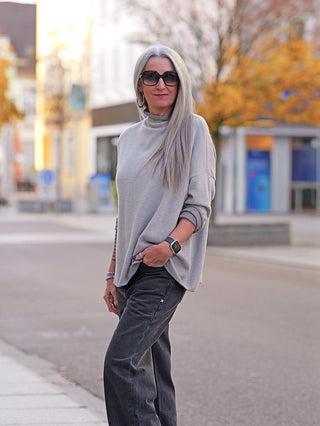 Frau auf leerer Straße trägt den OFF#DLY Pulli Siena beige, einen locker sitzenden, weichen Strickpullover mit leicht erhöhtem Kragen, kombiniert mit entspannten Jeans und Schmuck.