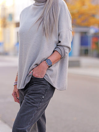 Person mit OFF#DLY Pulli Siena beige, locker sitzender Feinstrickpullover mit gerollten Ärmeln und kleinem Stehkragen, kombiniert mit dunkelgrauen Jeans und Schmuck, in urbaner Umgebung.