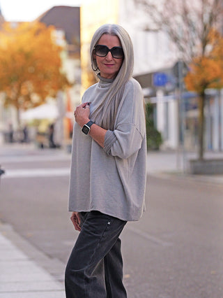 Person in einer ruhigen Stadtstraße trägt den OFF#DLY Pulli Siena beige mit überschnittenen Schultern, leichtem Stehkragen, kombiniert mit dunklen Hosen, Accessoires und einer Uhr, schafft einen lässigen Herbstlook.