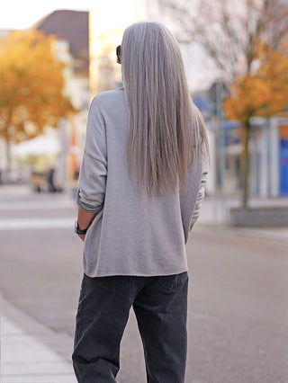 Person von hinten auf ruhiger Herbststraße, trägt OFF#DLY Pulli Siena beige mit fließender Oversize-Silhouette und leichtem Stehkragen, kombiniert mit lockeren Jeans und Sonnenbrille.
