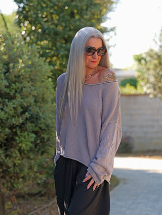 Frau trägt den OFF#DLY Pulli Abby grau aus Baumwolle, locker und off-shoulder gestylt, mit weitem Rundhalsausschnitt, kombiniert mit schwarzen Hosen, in lässiger, femininer Silhouette.