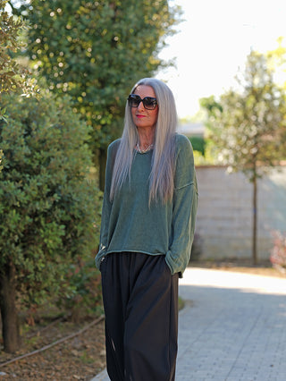 Mature Frau trägt OFF#DLY Pulli Abby green, locker geschnitten mit weitem Rundhalsausschnitt, kombiniert mit schwarzer Hose und silberner Kette, stehend auf einem Gartenweg mit Bäumen im Hintergrund.