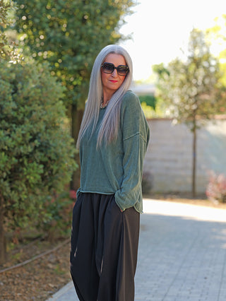 Person trägt den OFF#DLY Pulli Abby green, ein lässiger, weit geschnittener Pullover mit rundem Ausschnitt, ideal für einen entspannten Look, kombiniert mit schwarzen Hosen auf einem gepflasterten Weg.