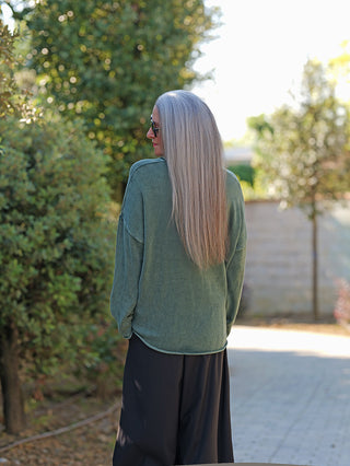 Frau in einem OFF#DLY Pulli Abby green mit weitem Rundhalsausschnitt und lockerer Passform, kombiniert mit weiten, schwarzen Hosen, steht auf einem gepflasterten Weg, umgeben von Bäumen.