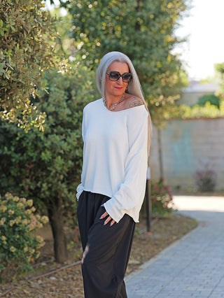 Eine Person trägt den OFF#DLY Pulli Abby white, ein locker sitzendes, off-shoulder Oberteil mit weitem Rundhalsausschnitt, kombiniert mit weiten schwarzen Hosen und silberner Halskette im Garten.