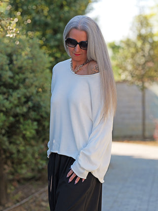 Frau trägt den OFF#DLY Pulli Abby white, ein weißes, locker sitzendes Baumwolloberteil mit weitem Rundhalsausschnitt, off-shoulder tragbar, kombiniert mit schwarzen weiten Hosen und einer goldfarbenen Kette.