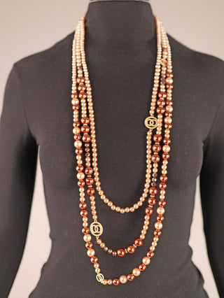 Die OFF#DLY Kunstperlenkette 3 Strang brown/gold hängt an einer schwarzen Schaufensterpuppe, bestehend aus langen Perlensträngen in champagner- und bronzefarbenen Tönen, mit goldfarbenen Logo-Medaillons.