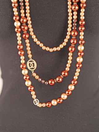 Die OFF#DLY Kunstperlenkette 3 Strang brown/gold zeigt lange, mehrlagige Perlenstränge in Kupferbraun, Champagner und Creme mit goldfarbenen Akzenten, elegant drapiert auf schwarzem Hintergrund.