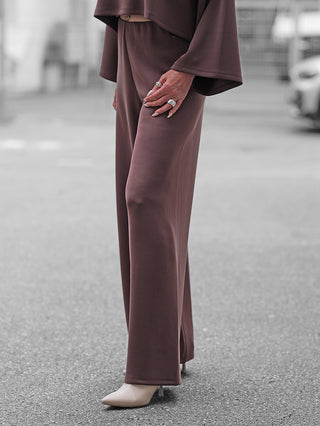 BSB Hose 212019 chokolate: Weite, schokobraune Hose mit fließendem Stoff, kombiniert mit spitzen, beigefarbenen Heels. Hand mit silbernen Ringen ruht am Oberschenkel, urbaner Hintergrund.