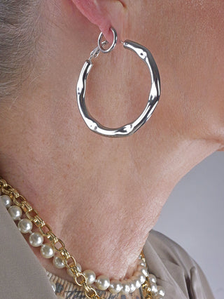 Nahaufnahme eines Halses mit OFF#DLY Creolen silber, einem großen, unregelmäßig geformten Silber-Ohrring im Ohr, kombiniert mit einer weißen Perlenkette und einer klobigen goldenen Halskette.