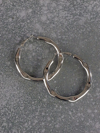 OFF#DLY Creolen silber, glänzende, silberfarbene Creolen mit leicht welliger, organischer Form, die auf einer grau gesprenkelten Oberfläche liegen. Durchmesser: 5 cm, aus antiallergischem Metall gefertigt.