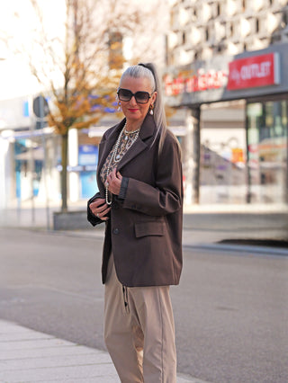 Frau trägt Lumina Blazer Schoko: Dunkelbrauner, oversized geschnittener Blazer mit klassischem Revers, sichtbar strukturierter Oberfläche und umschlagbaren Ärmelmanschetten, kombiniert mit Perlenketten und beigem Outfit.