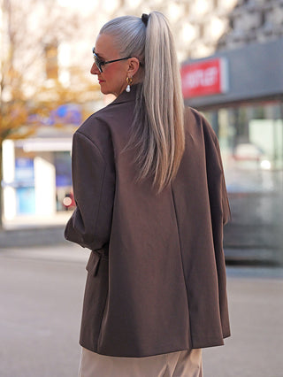 Person mit silbernem Pferdeschwanz trägt den Lumina Blazer Schoko in oversized, kombiniert mit hellem Hose. Blazer zeigt klassischen Revers und sichtbare Pattentaschen, perfekt für eleganten urbanen Stil.