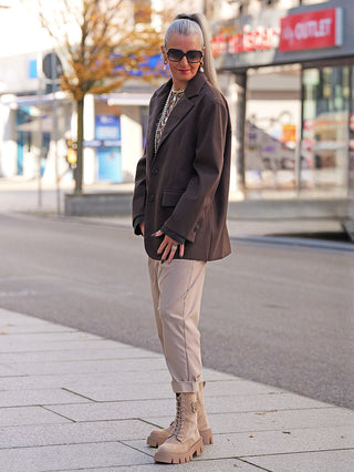 Erwachsener trägt den Lumina Blazer Schoko, oversized, mit klassischem Revers und variablen Manschetten. Kombiniert mit beigen Hosen und klobigen Stiefeln für einen smart-casual Look.