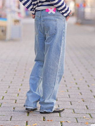 FDL Jeans Brooklyn Blue: Weit geschnittene, hellblaue Jeans aus 100 % Baumwolle mit auffälligem pinken Patch am Bund, kombiniert mit schwarzen, spitzen Schuhen auf einem gepflasterten Gehweg.