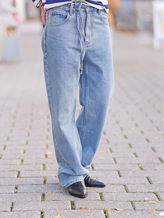 Die FDL Jeans Brooklyn Blue zeigt sich in einer hellen Waschung, mit weitem Schnitt und Bindegürtel, getragen auf einem Kopfsteinpflaster. Kombiniert mit schwarzen, spitzen Schuhen für urbanen Stil.