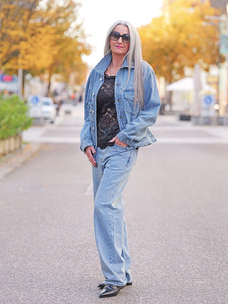 Frau trägt die FDL Denim Jacket Chicago blue, mit Oversize-Schnitt und ausgefransten Kanten, über schwarzem Spitzentop und passender Jeans, stehend auf einer herbstlichen, baumgesäumten Straße.