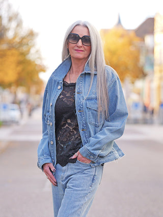 Frau mit silbernem Haar trägt FDL Denim Jacket Chicago blue in Oversize-Passform, erkennbar am markanten „X“-Stitching und ausgefransten Säumen, über schwarzem Spitzenoberteil und passender Jeans.