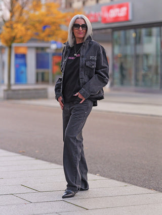 Person trägt die FDL Denim Jacket Chicago black mit oversized Schnitt, markantem „X“-Detail auf dem Rücken und ausgefransten Kanten, während sie auf einem Bürgersteig vor einem Geschäft steht.