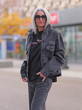 Ältere Frau mit Schulterlangem Silberhaar trägt die FDL Denim Jacket Chicago black, eine oversized Jeansjacke mit „FDL“-Stickerei auf der Brust, kombiniert mit dunklen Jeans für einen urbanen Look.
