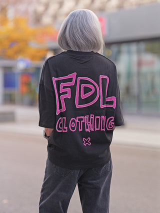 Person von hinten in urbaner Umgebung mit grauem Bob und schwarzem FDL Tee New York Graffiti, pinker Schriftzug FDL Clothing, lockerer Oversize-Schnitt, kombiniert mit dunklen Jeans.