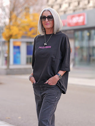 Frau mit grauen Haaren trägt das FDL Tee New York Graffiti black, ein schwarzes Oversize-T-Shirt mit pinken Graffiti-Prints. Kombiniert mit dunklen Jeans und Accessoires, verkörpert sie urbanen Streetstyle.