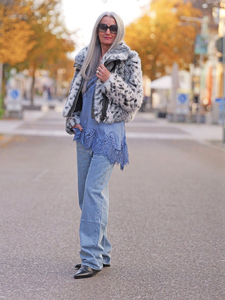 Frau trägt die OFF#DLY Fake Fur Jacke black/white mit Animal-Muster. Die Jacke hat einen kurzen Schnitt und kontrastierende Details, kombiniert mit einer blauen Tunika und Jeans für einen stilvollen Streetstyle-Look.