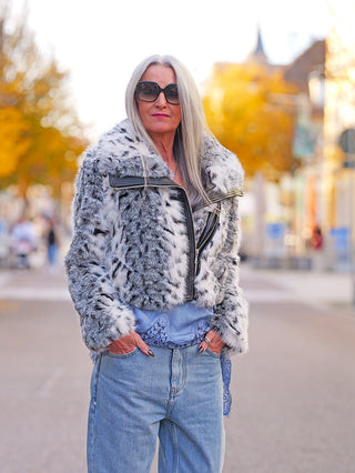 Eine ältere Frau trägt die OFF#DLY Fake Fur Jacke black/white, ein modisches Statement-Piece mit Animal-Muster und kontrastierenden Details, ideal für lässige Herbstlooks auf einer baumgesäumten Stadtstraße.