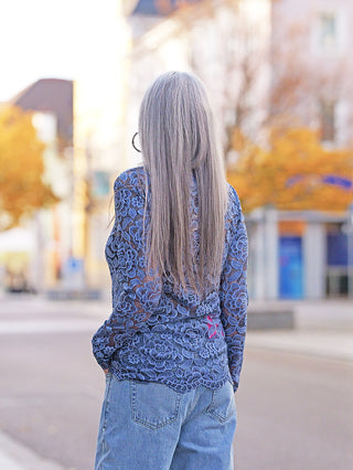 Person von hinten in einer Stadt, trägt das OFF#DLY Longsleeve Florenz blue, ein florales Spitzen-Oberteil mit langen Ärmeln, kombiniert mit blauen Jeans, umgeben von herbstlichem Stadtbild.