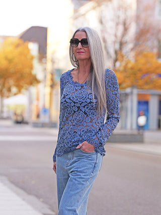Frau mit grauem Haar, trägt OFF#DLY Longsleeve Florenz blue mit feinem Spitzenmuster und figurbetontem Schnitt, kombiniert mit Jeans, während sie durch eine herbstliche Stadtlandschaft spaziert.
