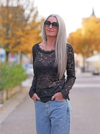 Person trägt das OFF#DLY Longsleeve Florenz black, ein langärmliges, schwarzes Spitzenoberteil mit figurbetontem Schnitt und rundem Halsausschnitt, während sie lässig auf einem herbstlichen Gehweg steht.