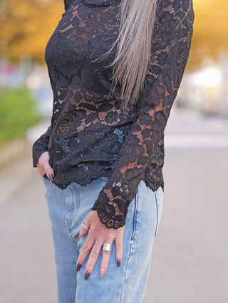 Person trägt das OFF#DLY Longsleeve Florenz black, ein figurbetontes, langärmliges Spitzentop, perfekt kombiniert mit hellblauen Jeans. Eleganz trifft auf lässige Moderne, ideal für stilbewusste Frauen.