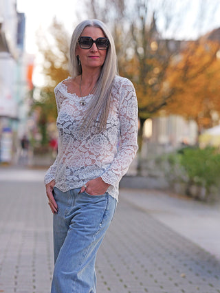 Frau trägt OFF#DLY Longsleeve Florenz, elegantes, weißes Spitzenoberteil mit langen Ärmeln, kombiniert mit blauen Jeans. Ideal für einen schicken, modernen Look.