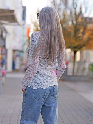 Person von hinten mit langen, grauen Haaren trägt das OFF#DLY Longsleeve Florenz white, ein Spitzentop mit floralem Muster, kombiniert mit blauer Jeans, spaziert auf Gehweg.