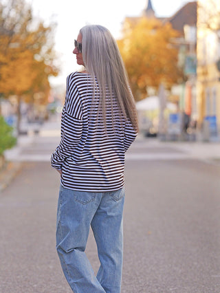Eine Person trägt das OFF#DLY Ringel Dora brown/white, ein langärmeliges, gestreiftes Shirt mit Rundhals und geschnittenen Kanten, während sie auf einer ruhigen Herbststraße spazieren geht.