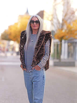Ältere Frau trägt die Off#DLY Fake Fur Weste Leo brown; auffällige Leopardenmuster-Weste über schwarz-weißem Streifenshirt, kombiniert mit Jeans, perfekt für einen modischen Herbst-Look.