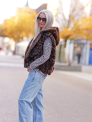 Person mit langen grauen Haaren trägt die Off#DLY Fake Fur Weste Leo brown, eine auffällige Leopardenmuster-Weste, kombiniert mit gestreiftem Oberteil und Jeans, auf einer ruhigen herbstlichen Straße.