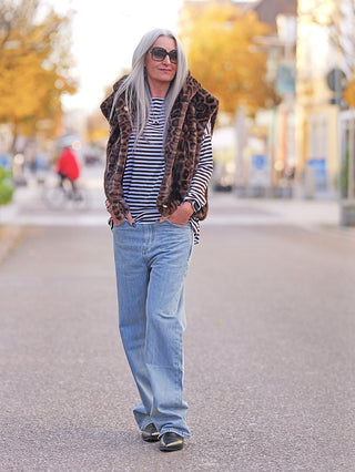 Ältere Frau trägt die Off#DLY Fake Fur Weste Leo brown, eine leopardenmusterige, weiche Kunstfellweste, über einem gestreiften Shirt, kombiniert mit Jeans, während sie durch eine herbstliche Stadtstraße spaziert.