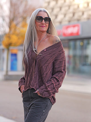 Person trägt die OFF#DLY West Ada braun, eine locker sitzende, mauve Strickweste mit V-Ausschnitt und Knopfleiste, über dunklen Jeans, kombiniert mit Sonnenbrille und großen Ohrringen.