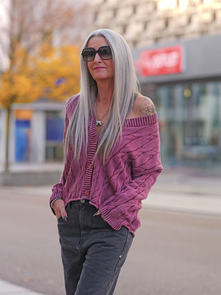 Eine Person trägt die OFF#DLY West Ada bordeaux, eine locker sitzende, kabelgestrickte Strickweste mit V-Ausschnitt, über dunklen Jeans. Ideal für lässige Streetstyle-Outfits.