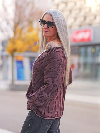 Frau trägt eine OFF#DLY West Ada braun, eine locker sitzende Strickweste mit markantem Strickmuster und V-Ausschnitt, über einem Schulter freien Pullover in urbaner Umgebung.