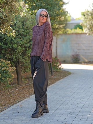 Person in einem losen, kabelgestrickten OFF#DLY Pulli Aida schoko, kombiniert mit weiten schwarzen Hosen und klobigen Schnürstiefeln, steht auf einem Gartenweg bei sanftem Sonnenlicht.