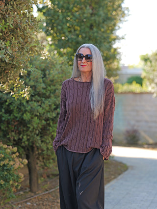 Ältere Frau trägt OFF#DLY Pulli Aida schoko, ein locker geschnittener, strukturierter Strick-Pullover mit betontem Ausschnitt. Kombiniert mit weitem, schwarzem Hosenbein auf gepflastertem Gartenweg stehend.