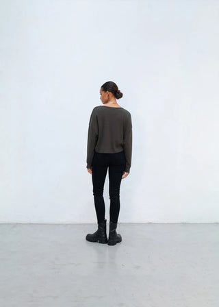 Person von hinten in einem minimalistischen Raum, trägt einen Outryght Kushi Pullover in lockerem Schnitt. Kombiniert mit schwarzen, engen Hosen und klobigen Stiefeln. Haare zum tiefen Dutt gebunden.