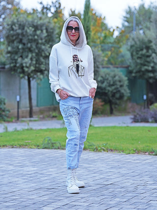 Frau in einem weißen OFF#DLY Sweat Women Hoodie mit Hut-Illustration, kombiniert mit hellen Jeans und weißen Stiefeln, steht lässig in einem Park auf Gehweg.