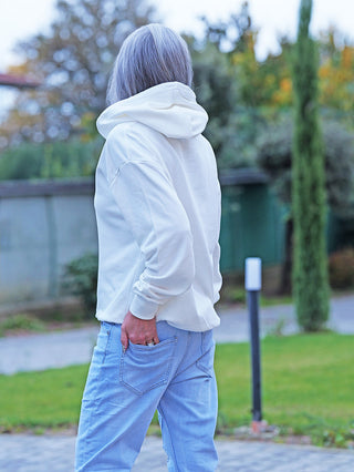 Eine Person trägt den OFF#DLY Sweat Women white Hoodie, lässig geschnitten mit Kapuze, auf einem Weg. Der Hoodie hat einen modischen Print auf der Vorderseite für einen stilvollen Akzent.