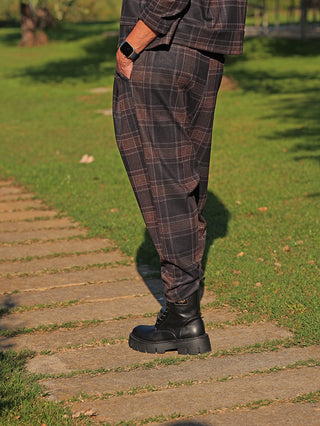 Person trägt OFF#DLY Pant Lilly kariert braun/grau, locker geschnittene Hose mit modernem Karomuster, kombiniert mit schwarzen Stiefeln auf Steinplatten im Freien. Smartwatch am Handgelenk sichtbar.