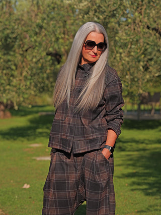 Die OFF#DLY Jacke Layla kariert braun/grau präsentiert sich mit einem modernen, boxy Schnitt und Stehkragen, getragen von einer Frau in einem parkähnlichen Umfeld, kombiniert mit passender Hose.