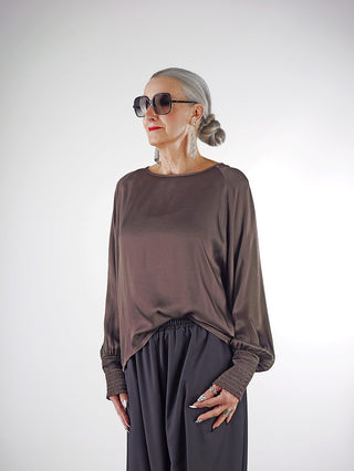 Die Frau trägt das OFF#DLY LS Shirt Valentina braun mit weiten Ärmeln und elastischen Bündchen, kombiniert mit weiten schwarzen Hosen für einen eleganten, minimalistischen Stil.